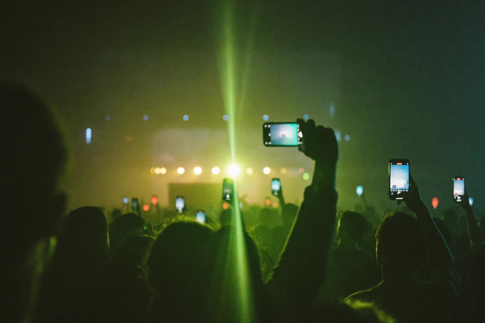 A concert crowd at night holding phones toward the stage, a lime-green light flare cutting the haze.