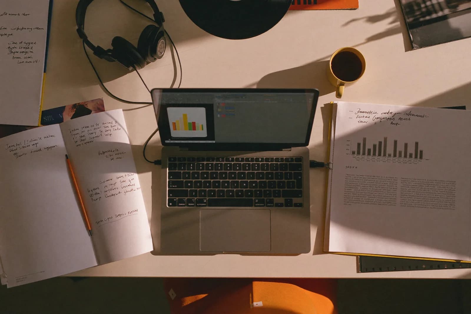 A desk in warm morning light with a laptop, coffee, printed charts, and headphones.