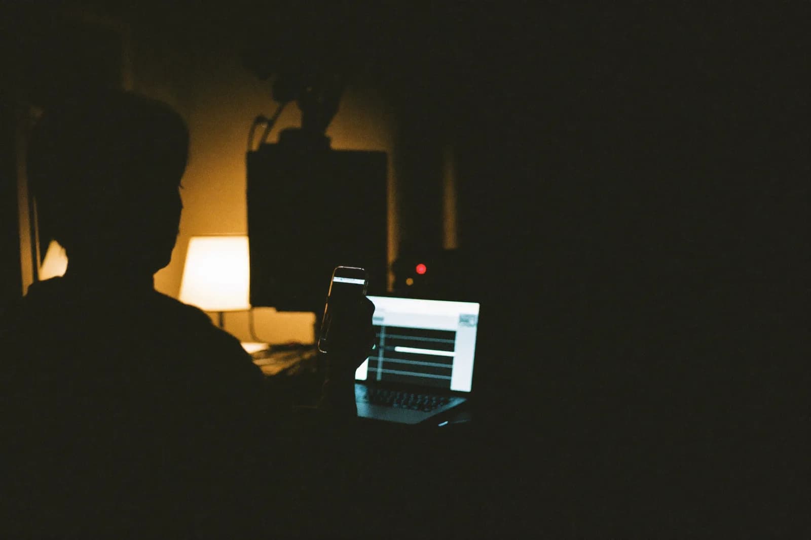 A musician alone in a dim home studio at night, phone in hand, laptop glowing beside them.